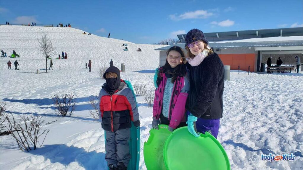 3 children stand with saucer sleds at the bottom of a snow covered sledding hill in the background with many children going down the hill 