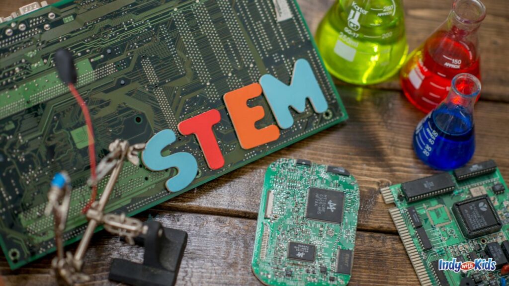 the word stem is laid out in colorful wooden letters on top of a computer motherboard. there are other computer chips lying around and three science jars with colored dye in each
