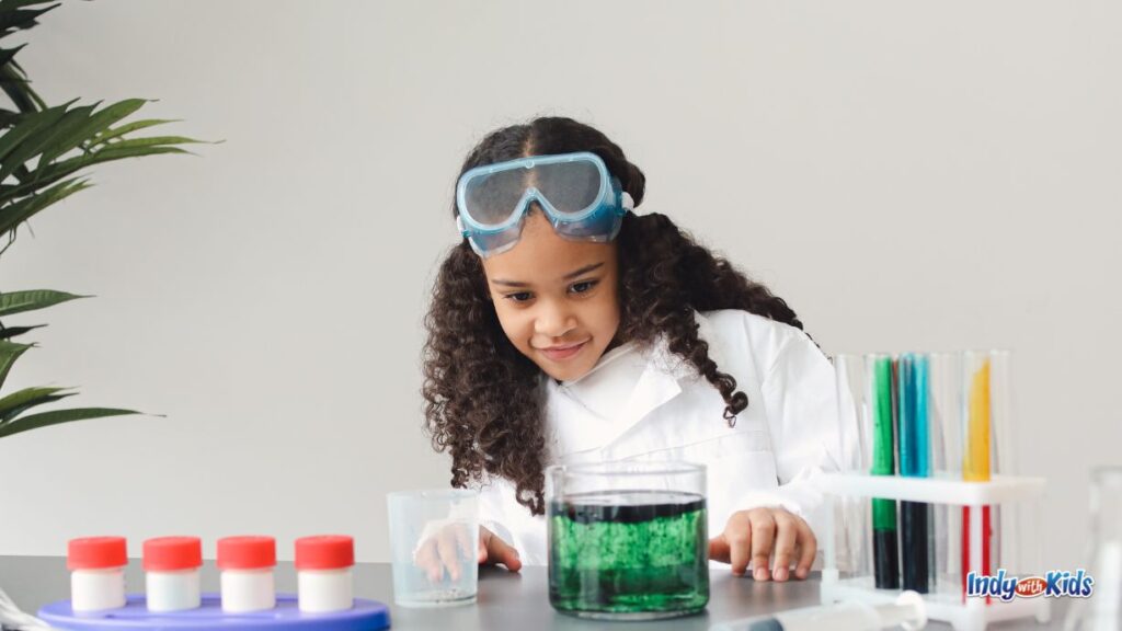 a school-aged girl is watching a science experiment in a glass jar with green oil in it in front of her. she has goggles on her forehead and there are vials and tubes of different colors on the table.