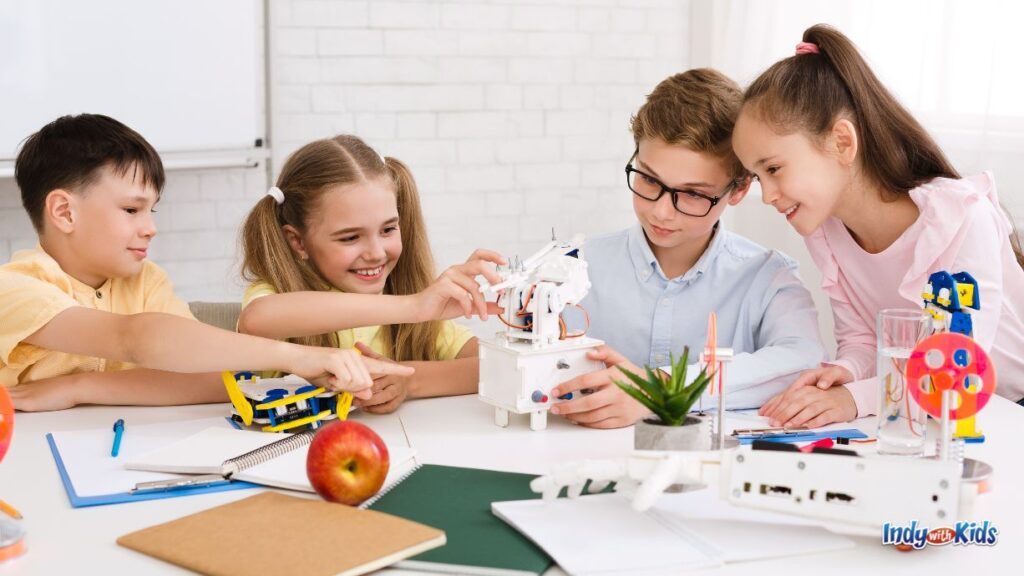 4 school-aged children sit at a table working with a white machine/robot they may have made or done coding with. there are other types of steam machines on the table