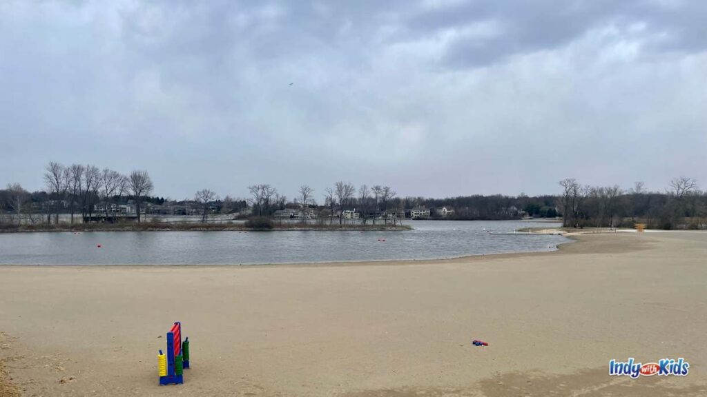 there is a large sandy beach next to a lake. there is giant connect four in the foreground and in the background behind the lake are trees.