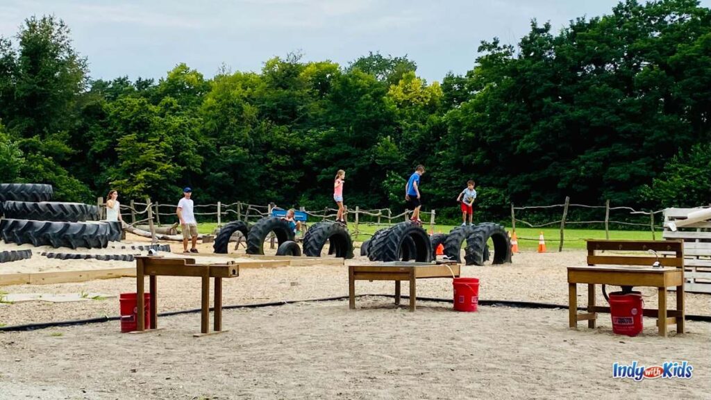 at fishers Agripark there is a large Sand Play area. in the foreground are mud kitchens with three sink stations. in the background there are tires to climb on and to jump from one to another.