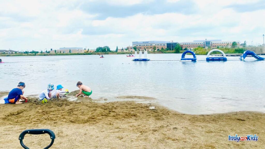 4 boys play in the Sand on a beach next to the water. in the middle of the lake is an inflatable obstacle course and behind that on the other side of the lake is a hospital building. there are also kayakers in the lake.