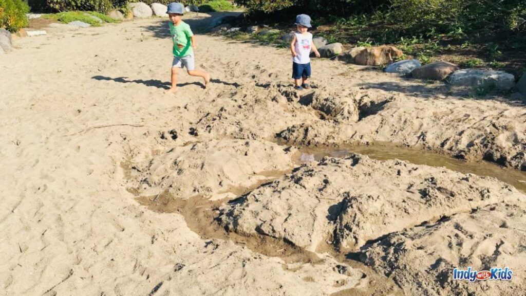 two little boys play in a large Sand Play area. there is a creek that they made filled with water.