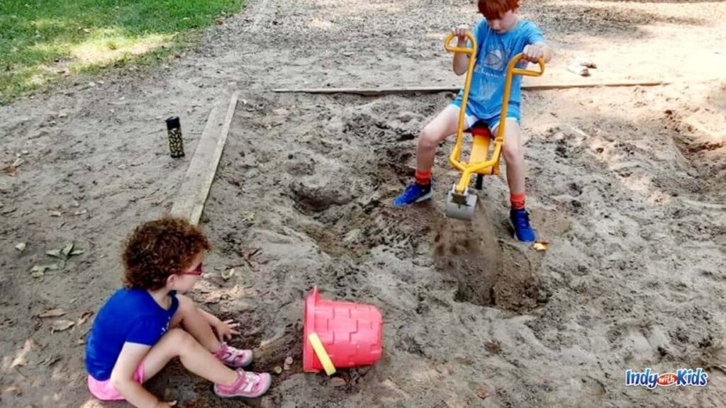 two children play in a sandbox. one is on a sitting digger and the other is on the ground with a red bucket.