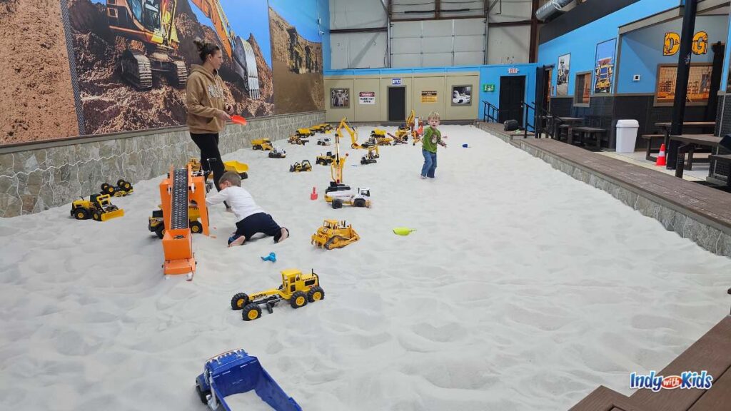 at an indoor Sand box there is a very large Play area filled with Sand, dump trucks, construction vehicles, and other toys. children are playing in the sand. there is seating on the side and a large digger mural on the wall.
