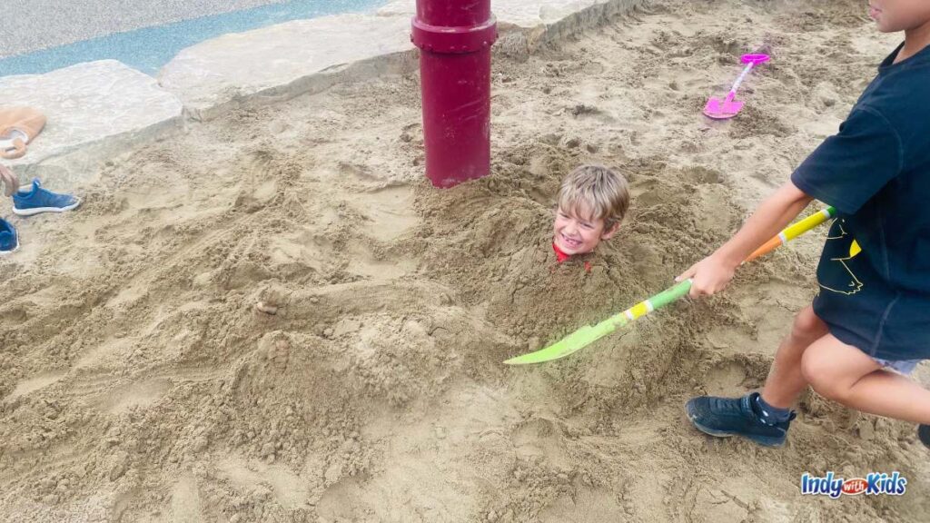 a child is buried in sand up to his head by another child holding a large plastic shovel