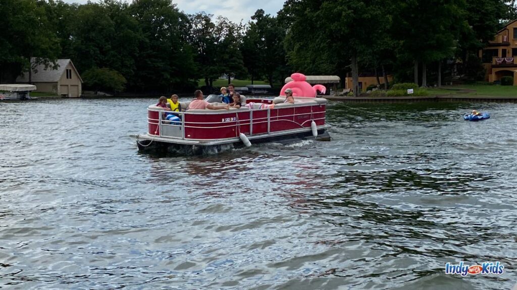 a red pontoon with several people in it cruises along on a lake with trees and homes in the background
