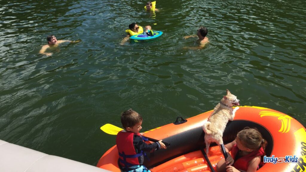several adults and children play in a lake. two children and a dog are in an inflatable boat. a baby floats in the water in a baby float with adults swimming next to him.