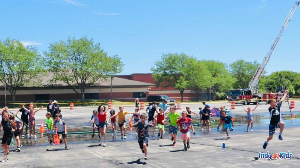 about 30 kids run and jump for joy in a parking lot at the Carmel Fire Department Summer Camp. there is a fire truck with its tall ladder extended in the background