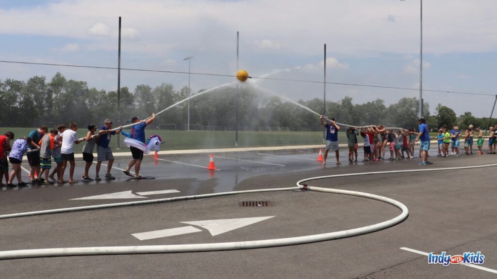 Carmel Fire Department Summer Camp: two groups hold a fire hose and each and are spraying a yellow ball on a wire in the air, trying to push the ball towards their opponets side