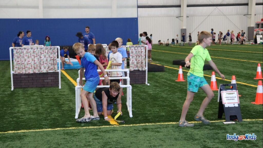 Carmel Fire Department Summer Camp: several children work through an obstacle course inside a building with astroturf. there are cones and plastic pipes set up as kids go under and through the obstacle