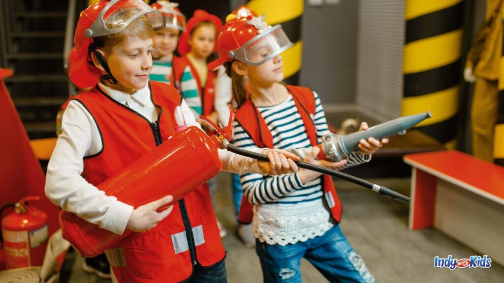 Carmel Fire Department Summer Camp: children dressed in red vests and red firefighter helmets hold up pretend fire gear