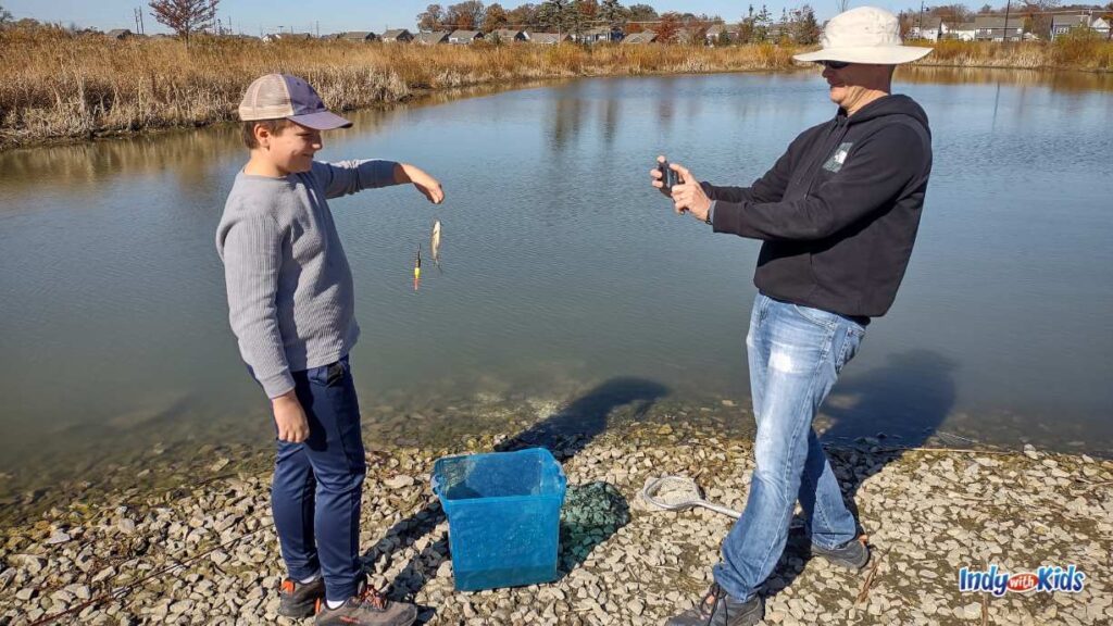 a father takes a photo of his son with a fish they just caught. there is a blue bucket on the ground and a lake behind them.