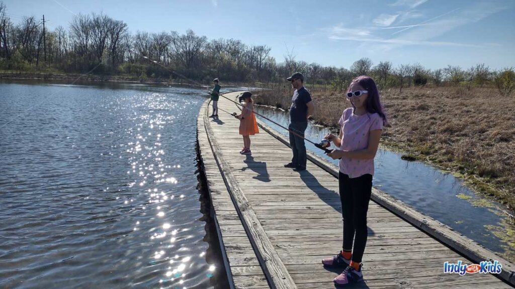 three children stand on a boardwalk with fishing nets cast into the lake on a sunny day. an adult male is watching over them.