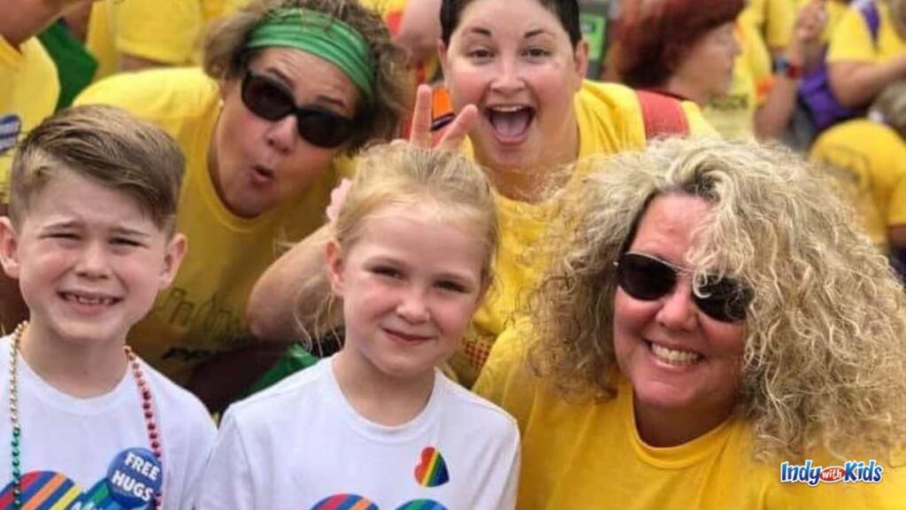 two children pose with three women at Indy pride. the children have white tshirts on and one has a pin that says free hugs. the three women behind them are all wearing yellow and one is giving the girl bunny ears.