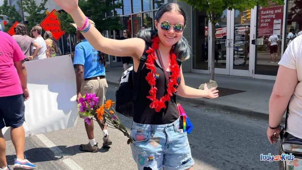 a young woman has her arms stretched out dancing/posing at the Indy pride parade. she has on a black tank top, a red flower lei, ripped jean shorts, and flowers with stems in her pocket.