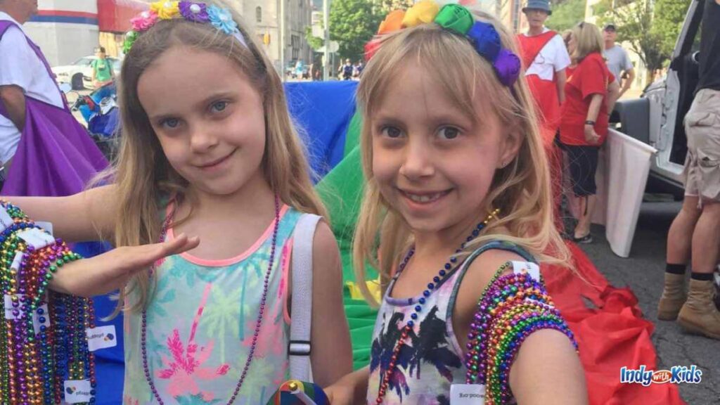 two little girls are all decked out at a festival with rainbow head bands and arms full of colorful beads.