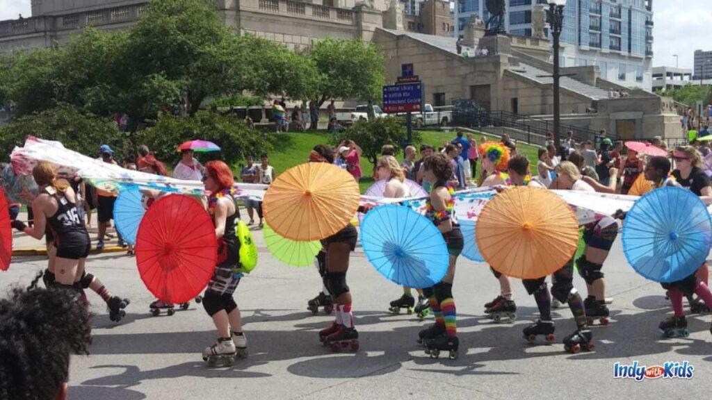 participants at the Indy pride parade roll down the route on rollerblades holding paper umbrellas of different colors in one hand and hold up a long sheet in between them in the other, much like a dragon at a parade.