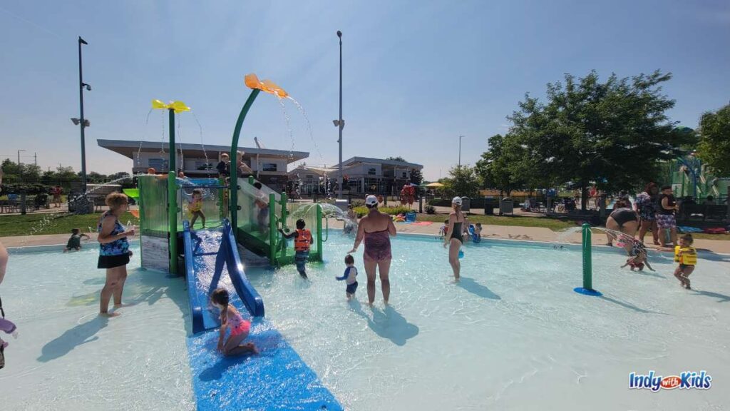 several adults and children play in the toddler pool at the Carmel Water park. adults wade in holding hands with babies and kids slide down the blue slide.