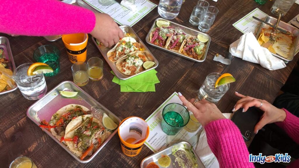 Carmel Indiana restaurants: several trays of tacos and drinks sit on a wooden table at social cantina. several arms wearing pink sweaters reach over the table to grab food.