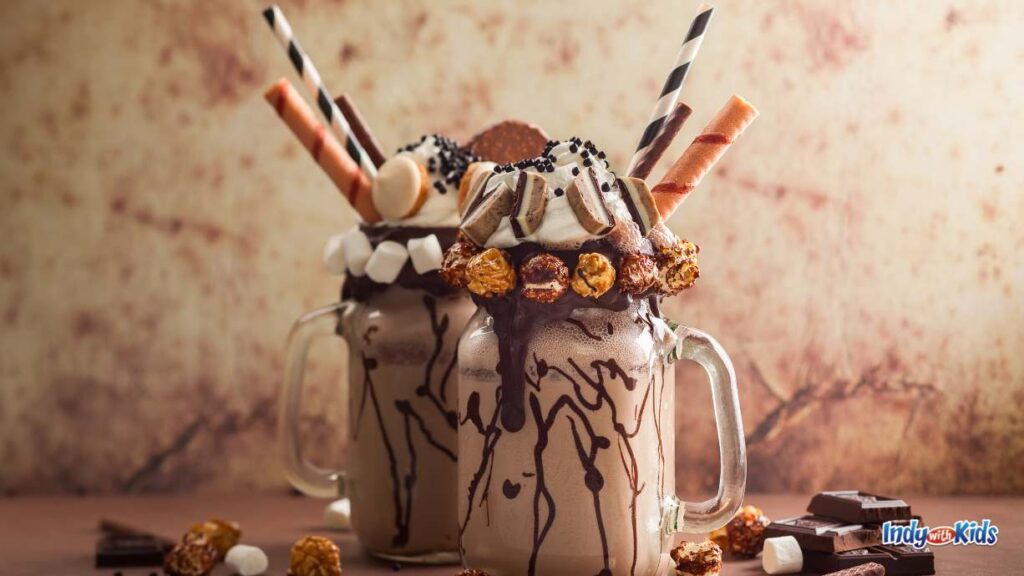 Spiked Shakes & Adult Slushies Near Me: two mason jars are filled with a chocolate shake and topped with mini marshmallows, candy, chocolate pieces, sprinkles, and three different types of straws (two edible). there are chocolate pieces on the table surrounding the mason jars.