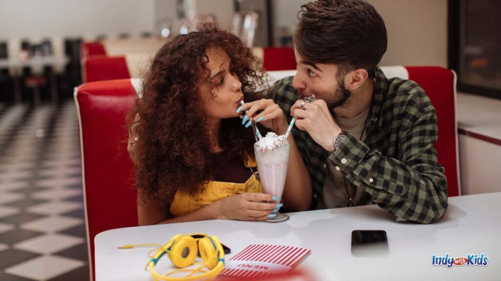 a couple on a date share a strawberry milkshake with two straws at a diner with red booths and black and white checkered floors