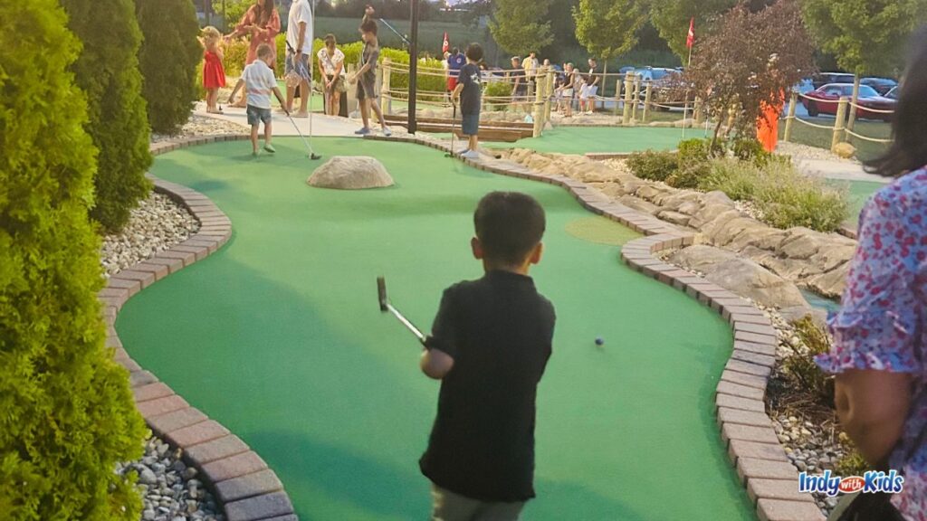 21 Best Westfield Indiana Restaurants | Family-friendly: a child has just hit a golf ball at a putt putt course. there are several children on the green at the other end near the hole.