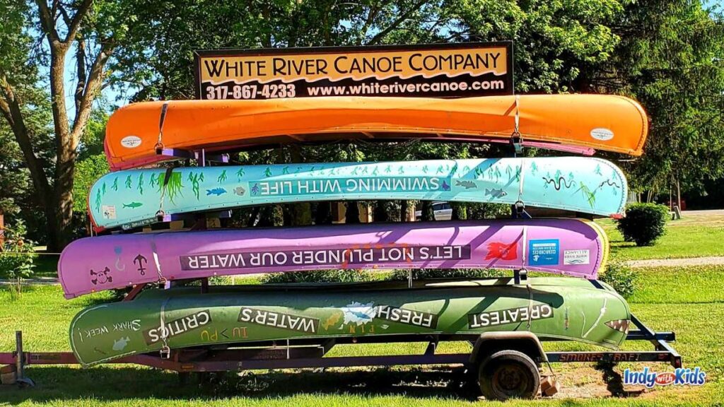 signage for the white river canoe company consists of four canoes turned upside down one on top of another. all different colors with different phrases on each. the green says greatest fresh water's critics. purple says let's not plunder our water. the blue one says swimming with life. the orange on top is plain orange.