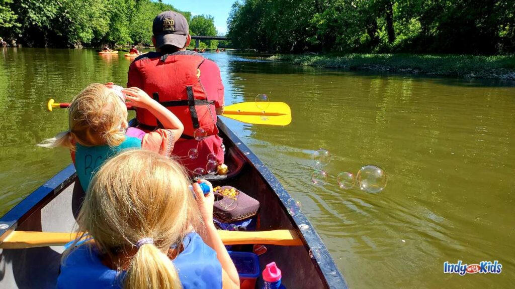 a man paddles a canoe wearing a red life jacket and a backwards blue baseball hat. his two daughters ride behind him in the canoe and are blowing bubbles. there are other canoers in front of him. the water is brown and there are trees on the banks on either side of the river.