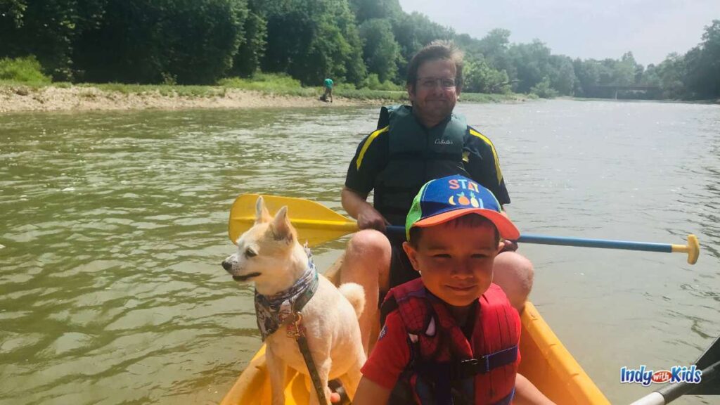 a man sits in a yellow kayak with a small white dog and a little boy with a stay cool hat and a red life jacket on in front of him. they are on a river and you can see a person on the river bank behind them.