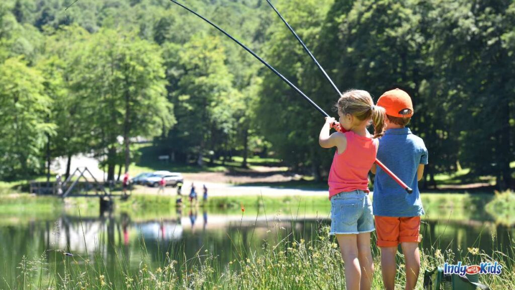 where to go fishing near me: a little boy and girl hold large fishing poles over a lake. they are faced away from the camera facing the lake. you can see cars, people, and playground equipment in the distance on the other side of the lake.