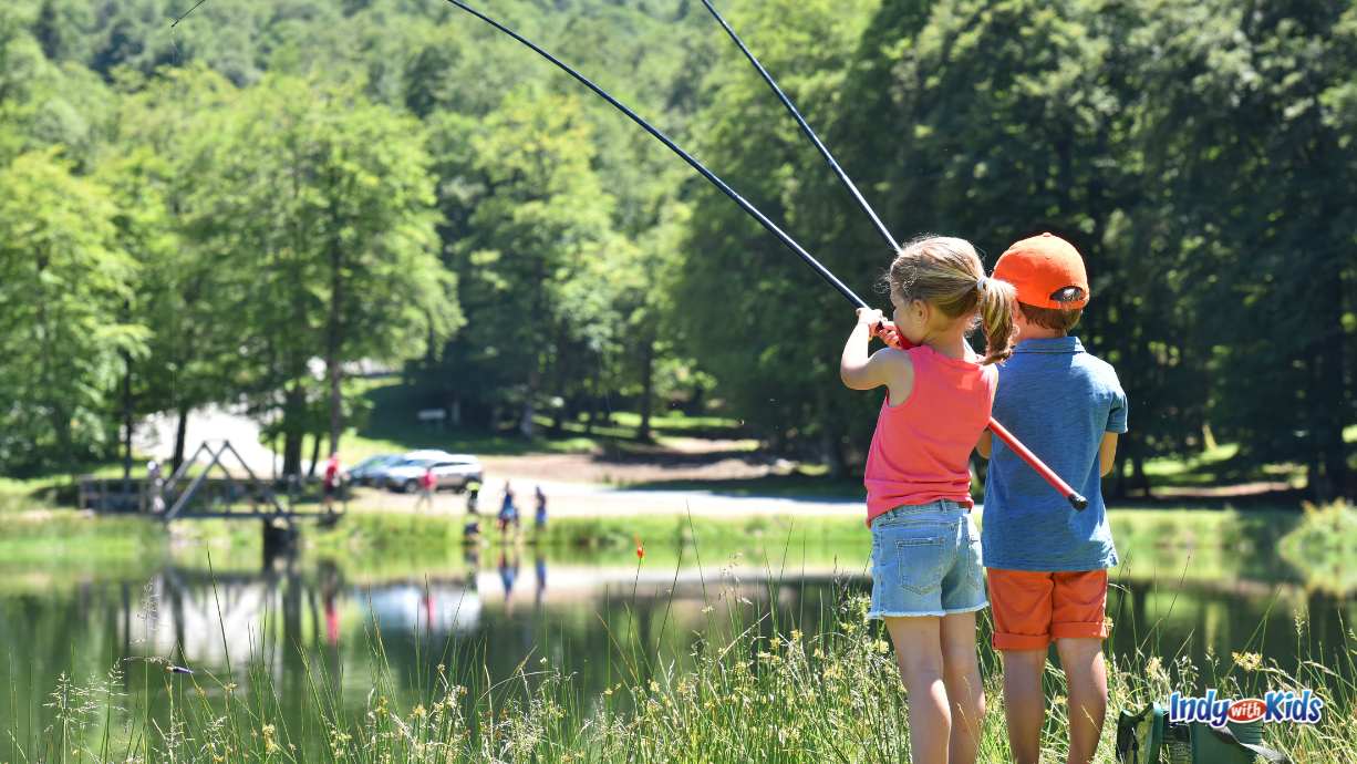 Two children stand at the edge of a quiet pond holding fishing poles on a sunny day, surrounded by green trees and grass at a family-friendly fishing spot.