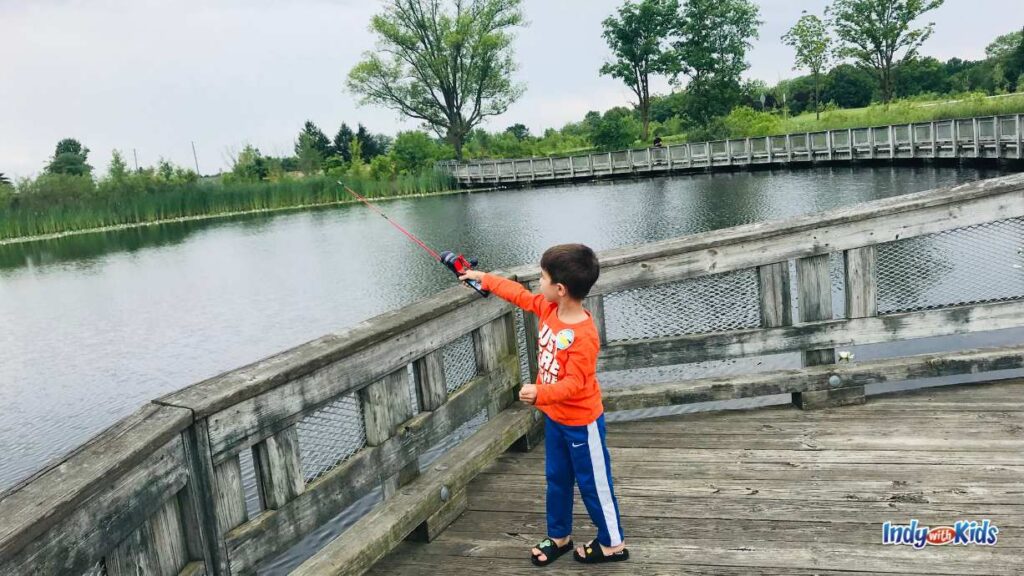 where to go fishing near me: a little boy wearing an orange shirt and blue pants stands on a wooden pier at west Park. he is holding a fishing pole over the railing and looking out over the pond.