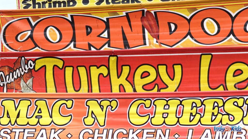state fair food recipes: an orange sign with yellow letter reads, in different fonts each, corn dogs, turkey legs, Mac n cheese. shrimp, steak chicken, lamb.
