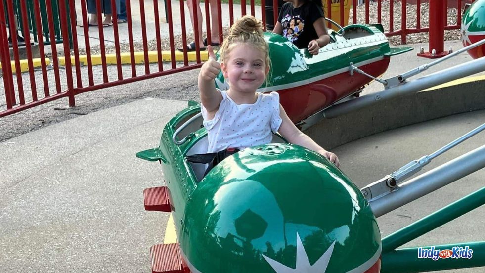 A child gives a thumbs up while seated on a ride at a theme park.