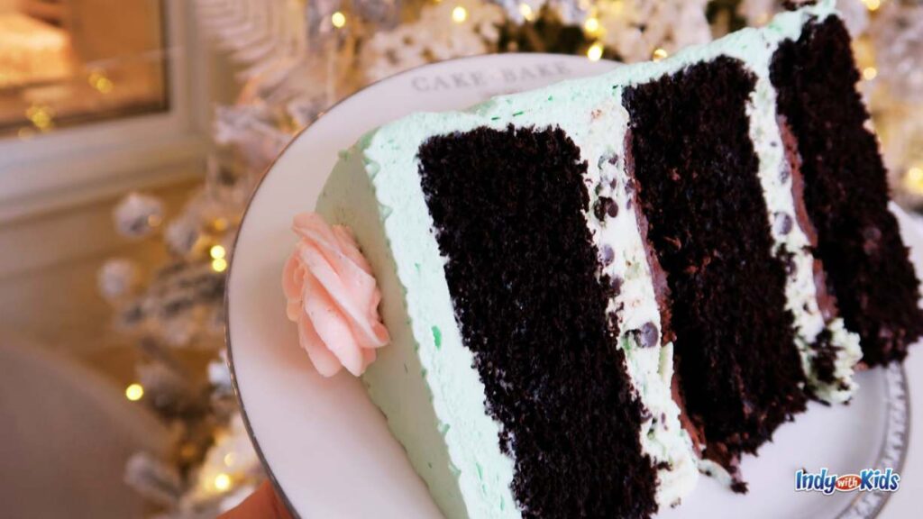 a giant slice of cake bake shop cake on a plate in front of a white frosted christmas tree. the Cake is three layer chocolate with mint frosting.