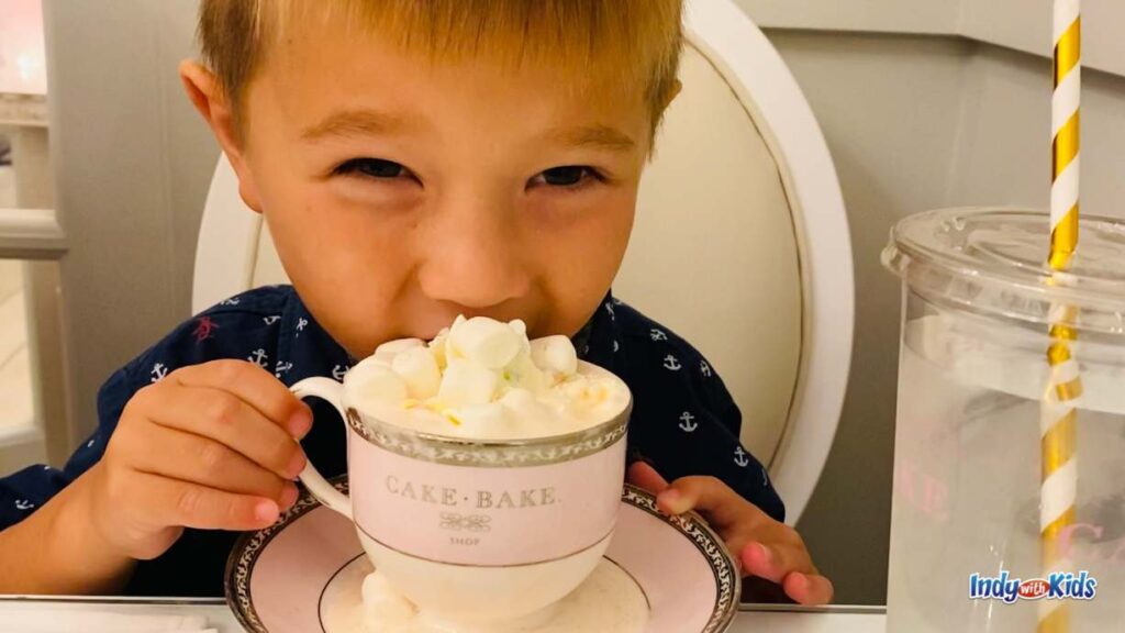 a little boy in a blue shirt sips on a unicorn hot chocolate from a teacup and saucer. the hot chocolate is topped with mini marshmallows.