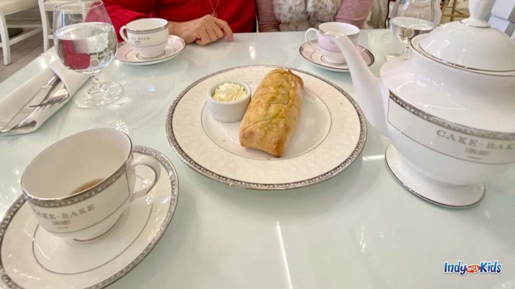 a table is filled with teacups on saucers, a bread and butter plate, waters, and a large cake bake shop teapot.