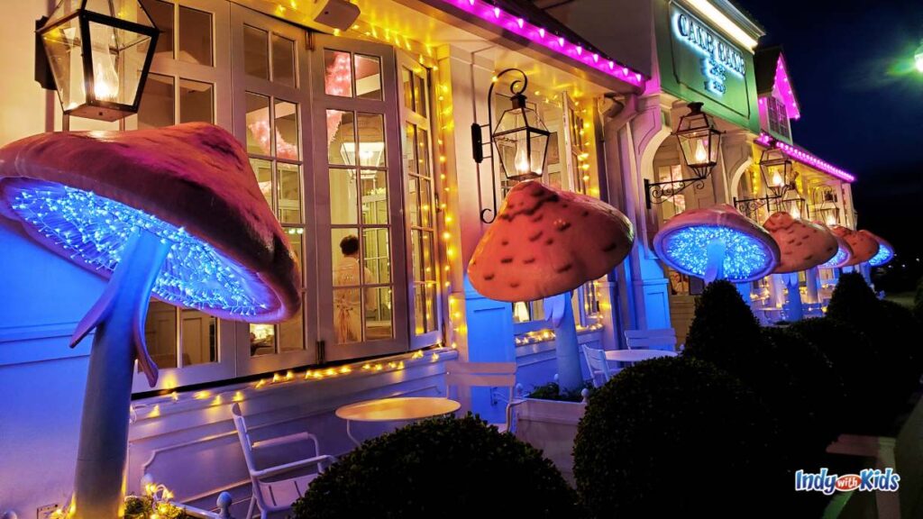 the Cake bake shop at night. the facade is lit up with yellow and purple string lights. the are large felt mushrooms in planter boxes lit up as well.