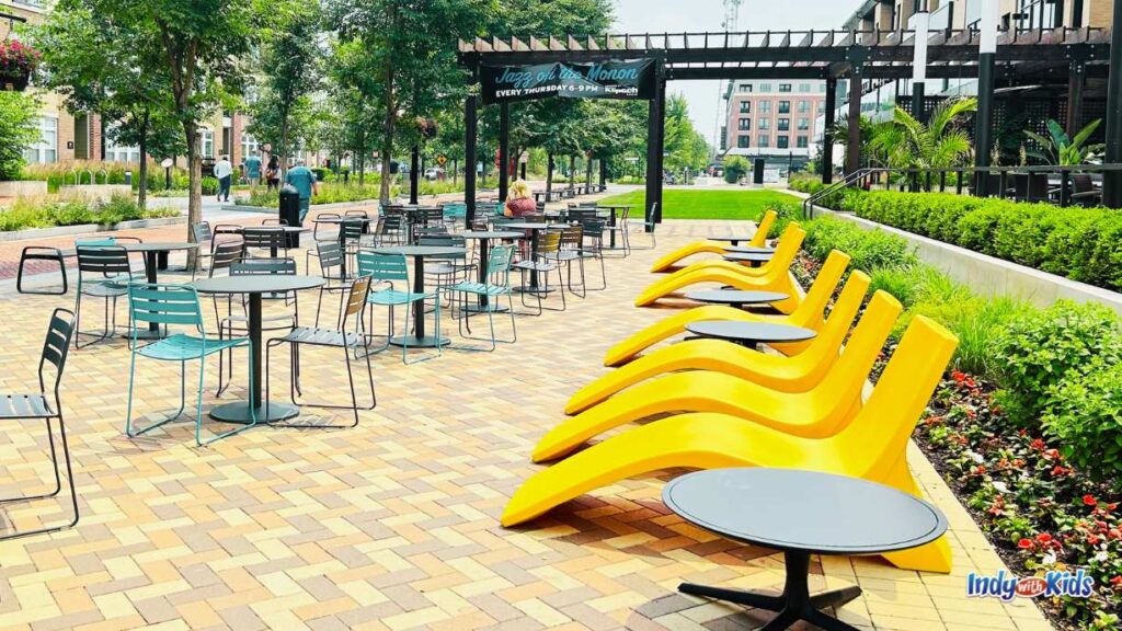 a view of seating areas at the Carmel Arts & Design District a long the monon trail. there are metal chairs and tables to the left and yellow plastic sunbeds on the right. 