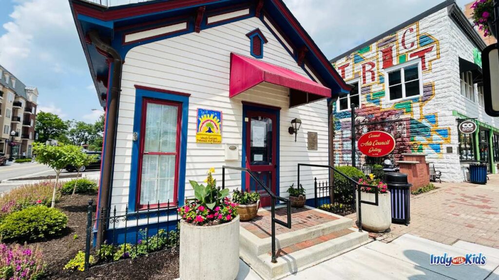 a tiny one room house is the home to the world's smallest children's art gallery. there is a red awning over the door and a garden surrounding the house. 