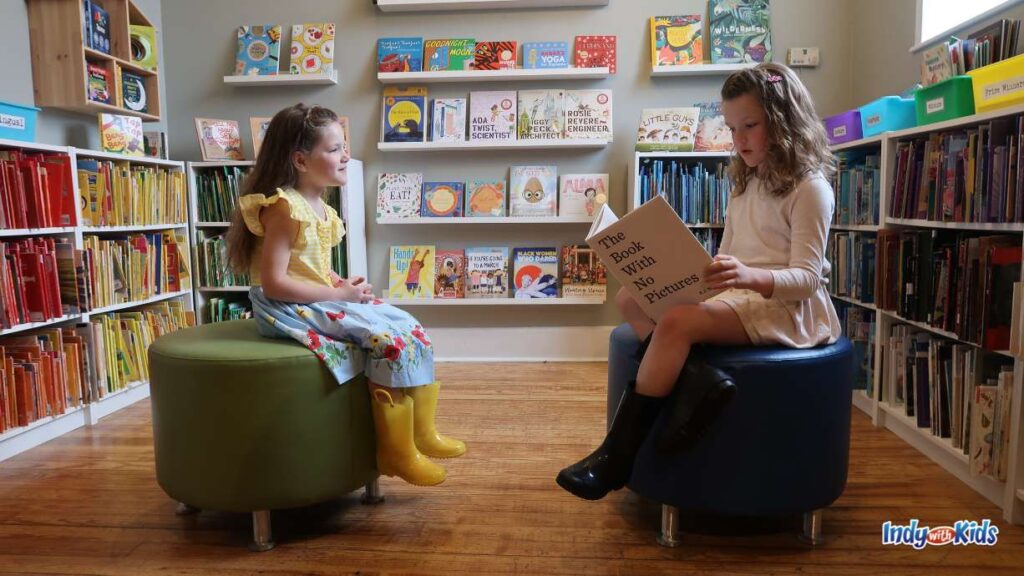 things to do in august: an older girl reads to a younger girl at a bookstore. they are both sitting on ottomans.