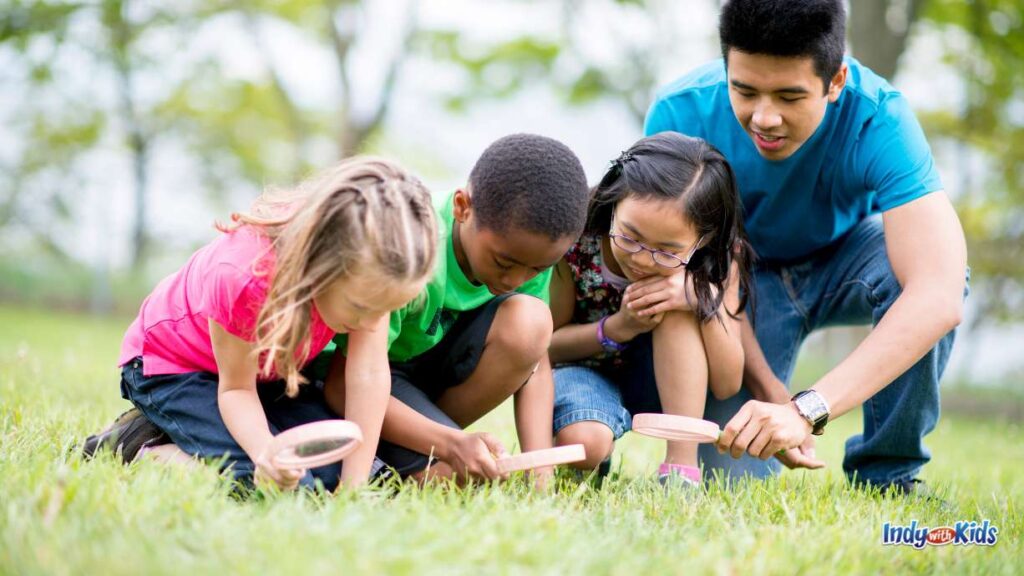 things to do in august: three kids and an adult inspect insects with magnifying glass outside in the grass