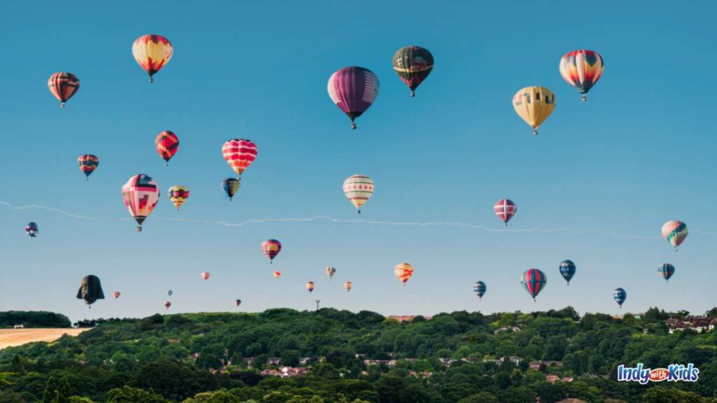 Several dozen hot air balloons fill a clear sky over trees and houses.