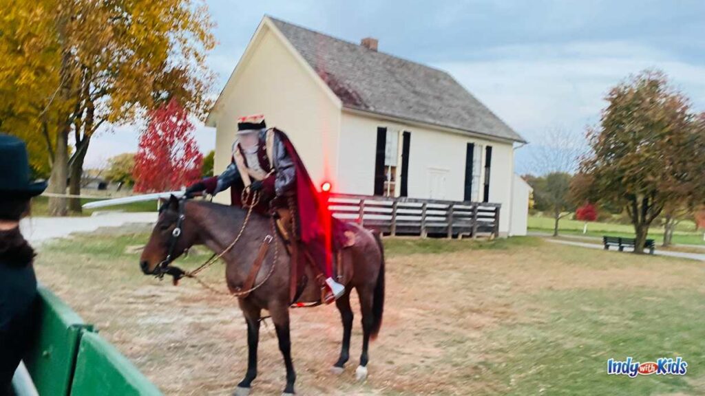 a headless horseman on a brown horse rides up next to a green wagon at the Headless Horseman Festival at Conner Prairie.