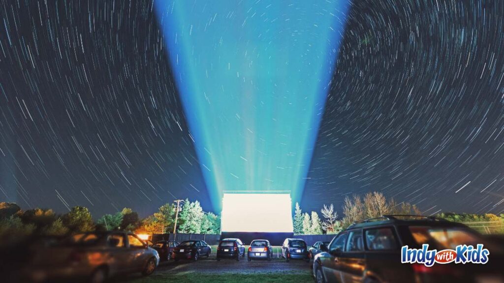 Best Drive In Movie Theater Indianapolis Area: cars are parked watching a movie at the drive in under the stars. the projection screen shines and lights up the sky above it.