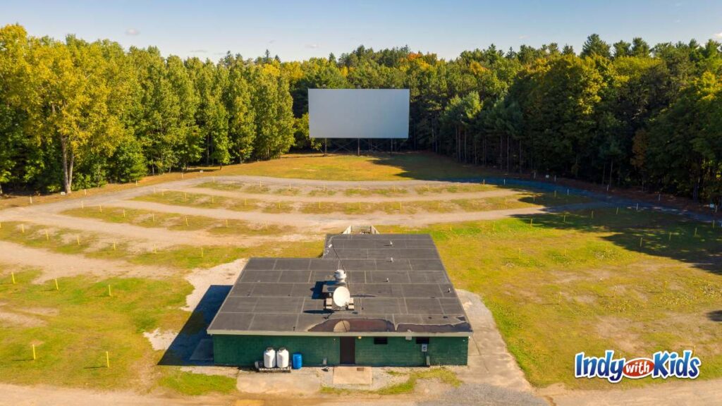 Best Drive In Movie Theater Indianapolis Area: a drive in movie theater sits in a huge grassy field surrounded by large trees and woods. the large projection screen is in the middle back and the concessions stand is in the forefront.