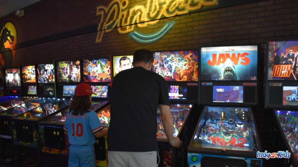 A man and a child in a baseball hat and outfit play on pinball machines next to each other at Boss Battle Games