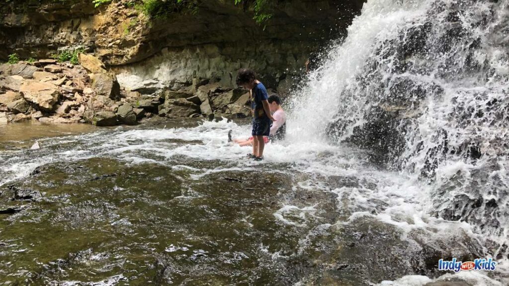 13 Fascinating Waterfalls in Indiana: two children play at the base of a waterfall, one sitting and one with his back to the waterfall